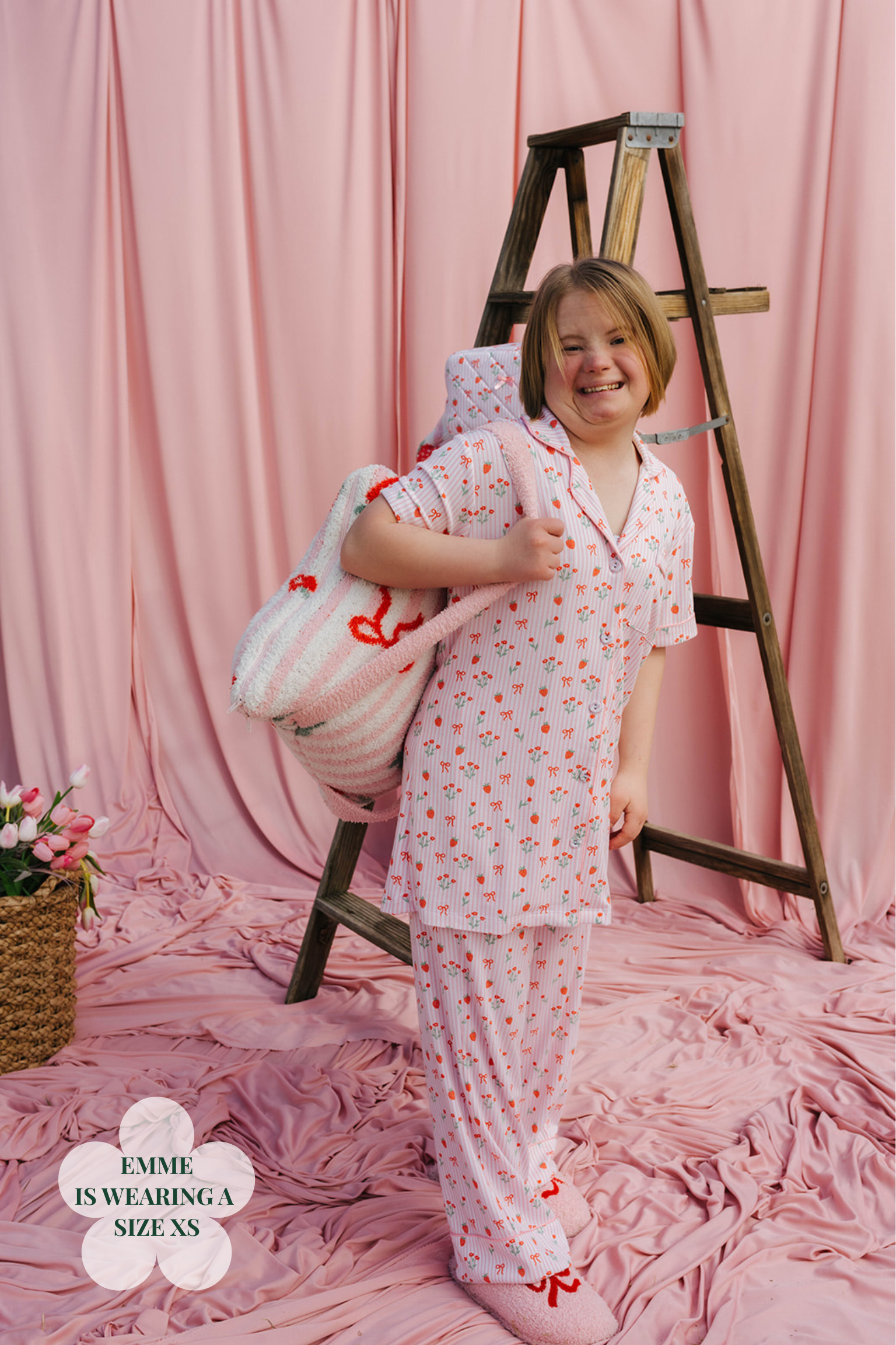 Child wearing patterned pjs smiling next to a wooden ladder against a pink curtain backdrop.