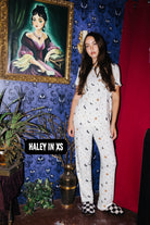 A woman stands in a styled room wearing a white short-sleeve pajama set with small black and gold motifs.