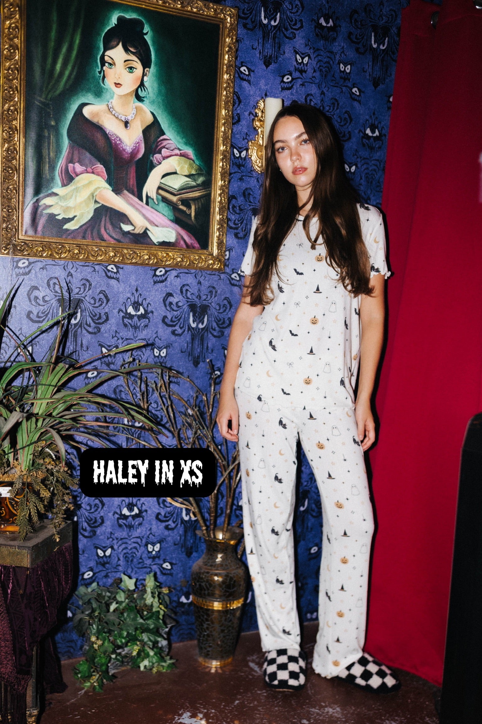 A woman stands in a styled room wearing a white short-sleeve pajama set with small black and gold motifs.