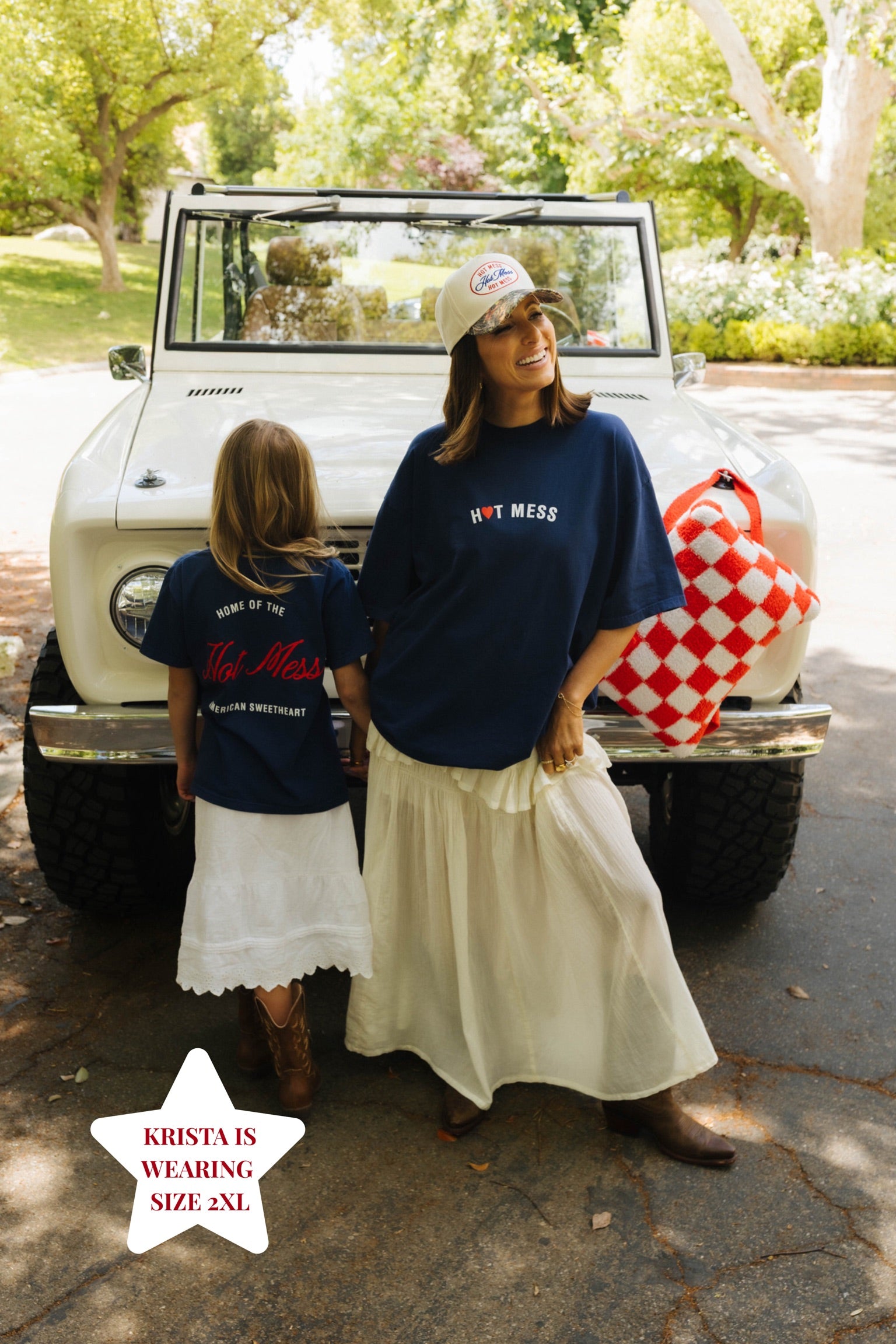 Two women standing in front of a vintage truck with a checkered blanket, wearing navy blue shirts with text.