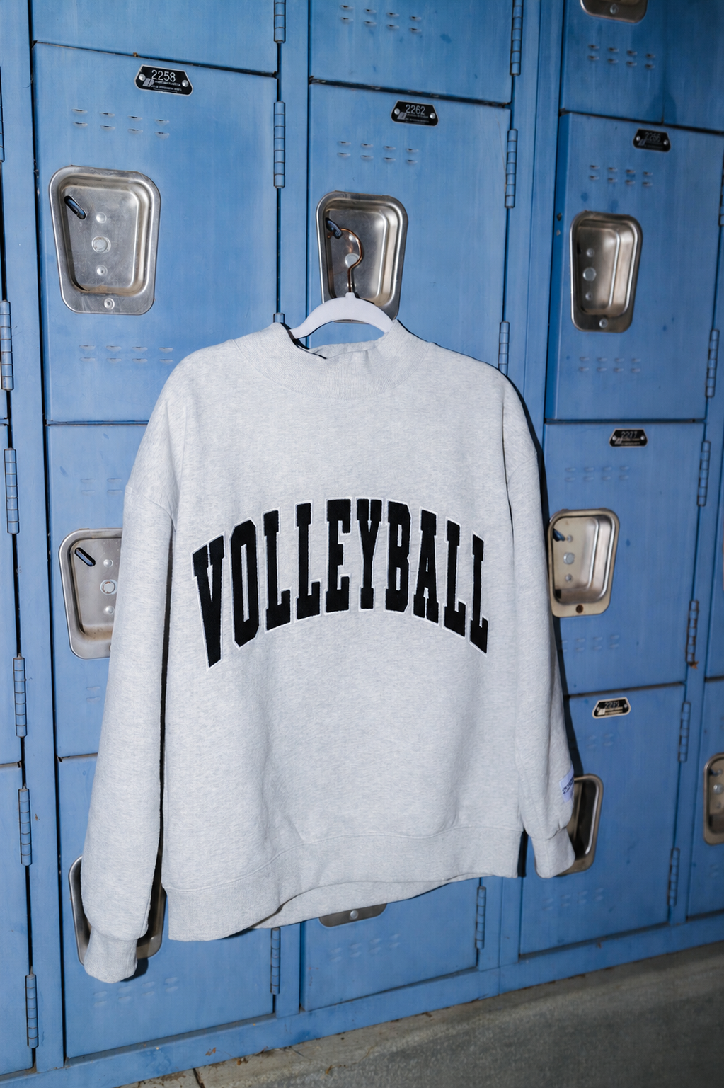 A light gray heather crewneck sweatshirt with 'VOLLEYBALL' printed in black arched block letters across the chest, hanging on a white hanger in front of blue school lockers.