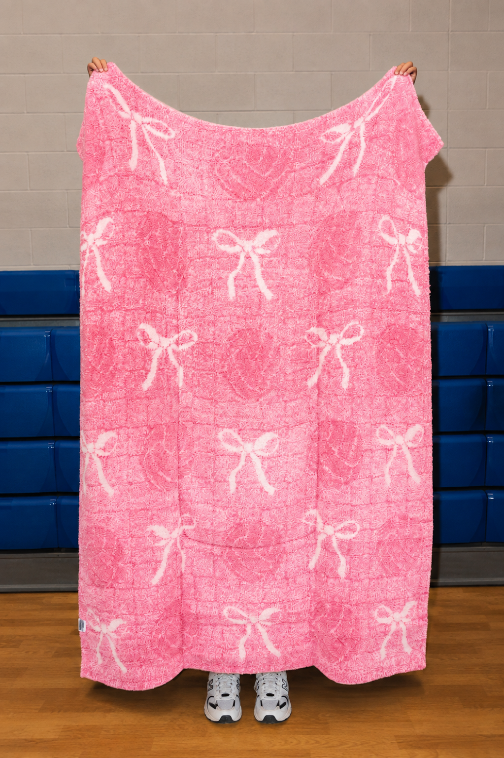 A pink blanket with a white bow and heart pattern is held up by a person in a gym with blue padding on the wall.