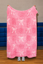 A pink blanket with a white bow and heart pattern is held up by a person in a gym with blue padding on the wall.