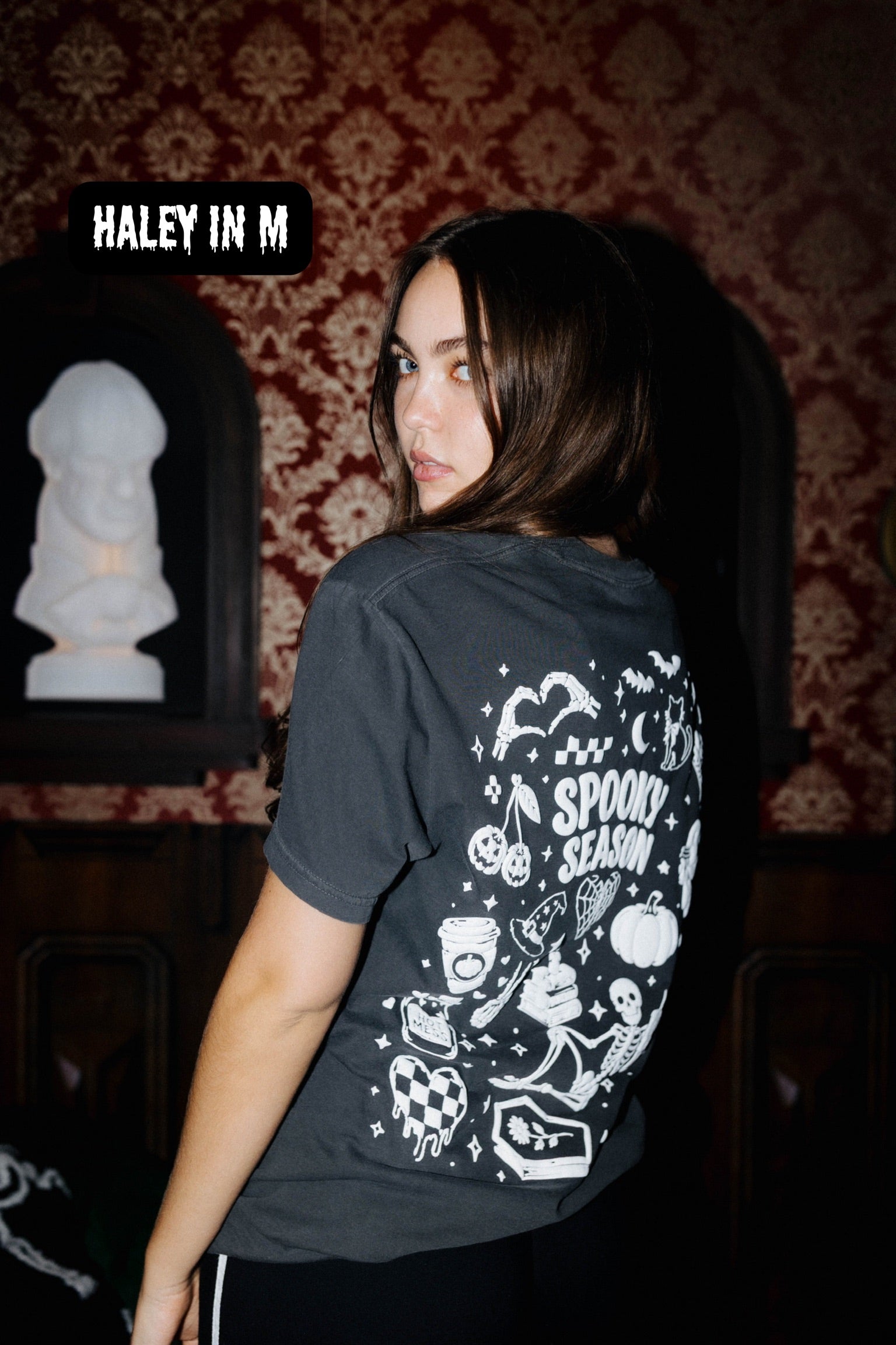A woman wearing a dark gray graphic t-shirt with a white 'Spooky Season' print on the back, standing in a dimly lit room with patterned wallpaper.
