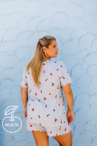 A woman wearing a light blue and white strawberry-print short-sleeve top and matching shorts, photographed from the back against a textured light blue wall.