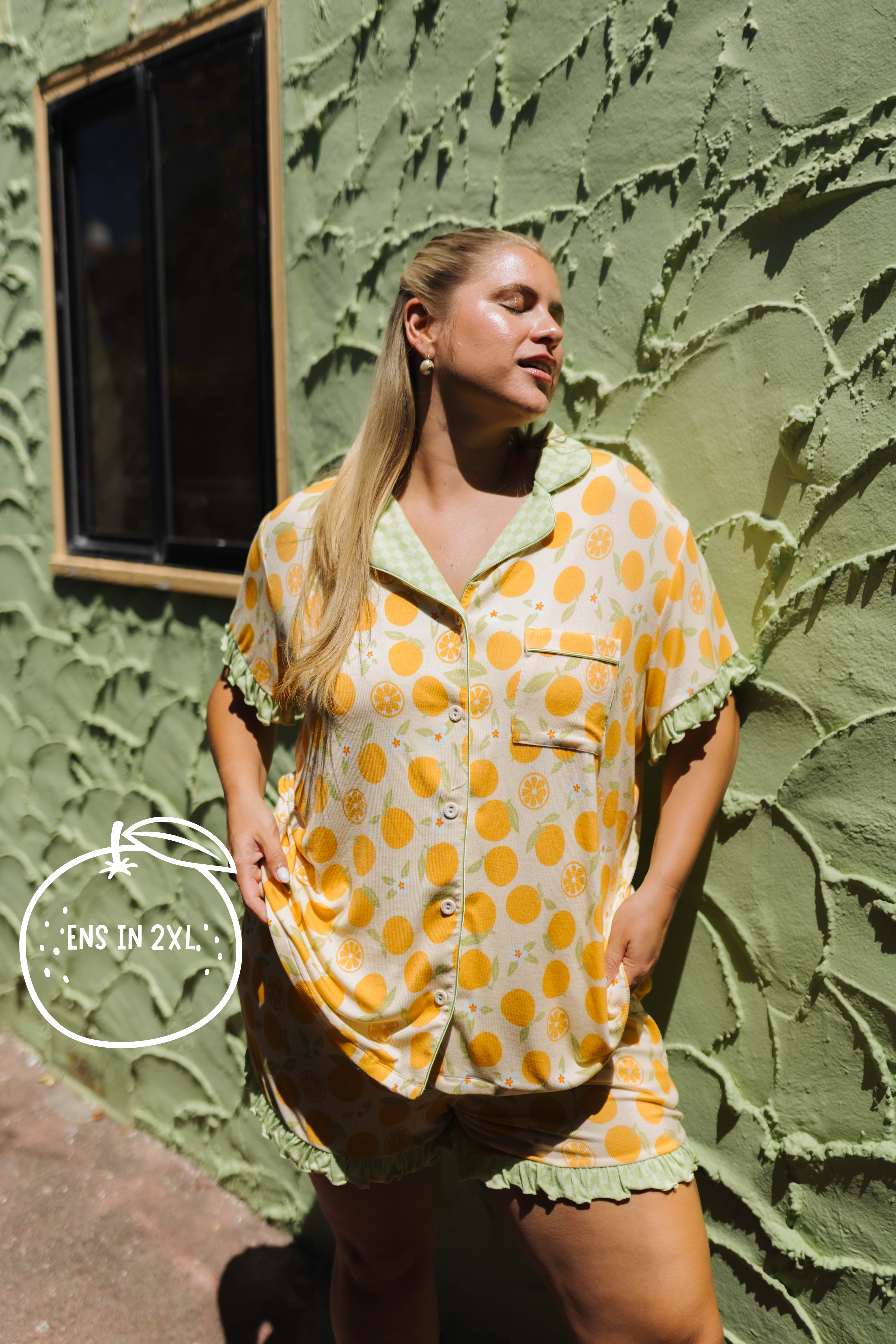 A woman modeling an orange citrus-patterned short-sleeve pajama set with green ruffled trim, standing against a textured olive green stucco wall.