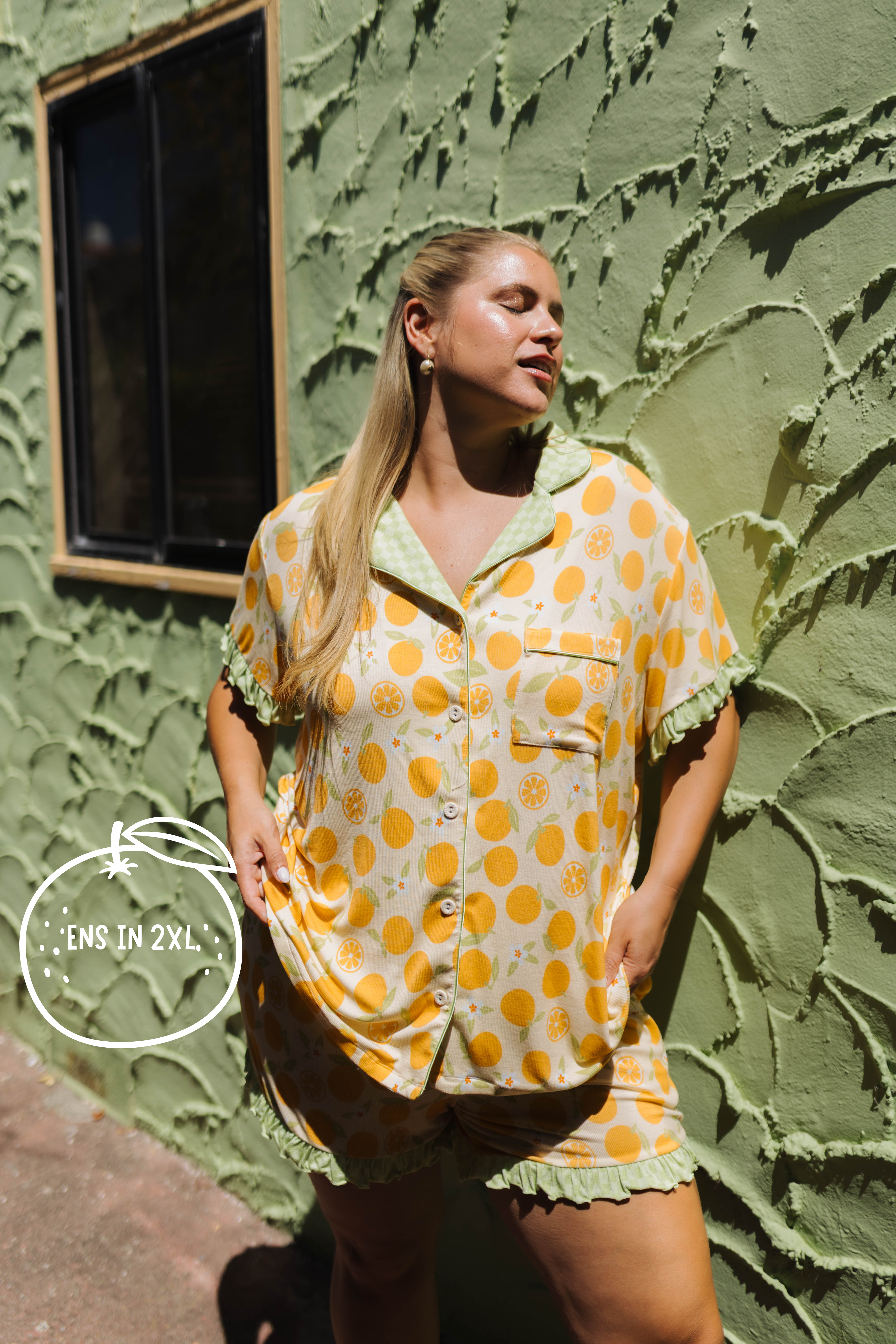 A woman modeling an orange citrus-patterned short-sleeve pajama set with green ruffled trim, standing against a textured olive green stucco wall.