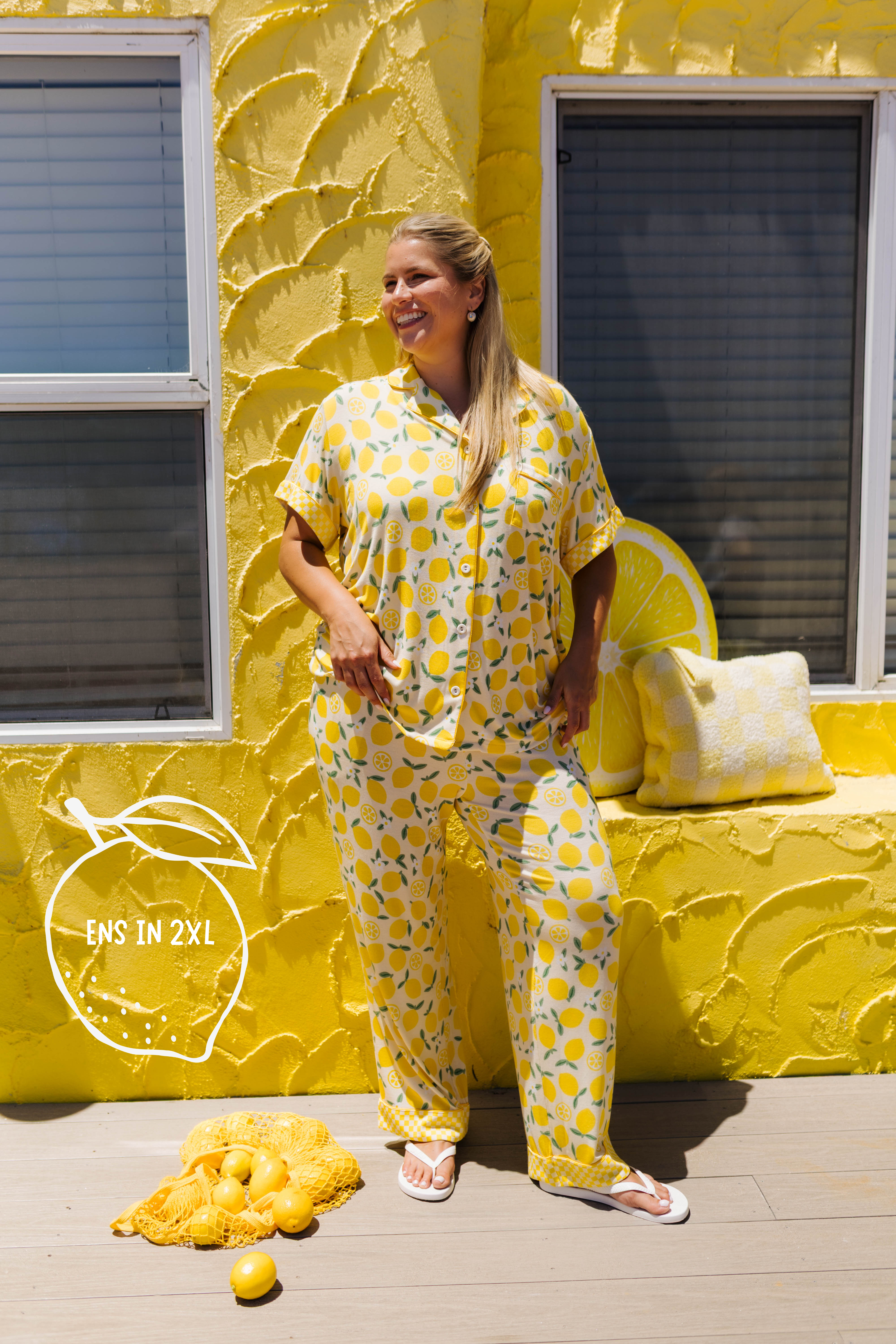 A woman modeling a yellow lemon-patterned pajama set, standing in front of a textured yellow wall decorated with lemons and pillows.
