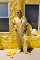 A woman modeling a yellow lemon-patterned pajama set, standing in front of a textured yellow wall decorated with lemons and pillows.