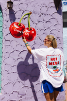 A woman wearing a white graphic t-shirt and blue shorts stands in front of a textured lavender wall, reaching toward a large 3D cherry mural painted on the wall. The t-shirt features a large graphic on the back with the text 'HOT MESS' and 'Ensley in 2XL'.