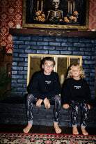 Two children wearing black Spooky Season sweatshirts by a fireplace