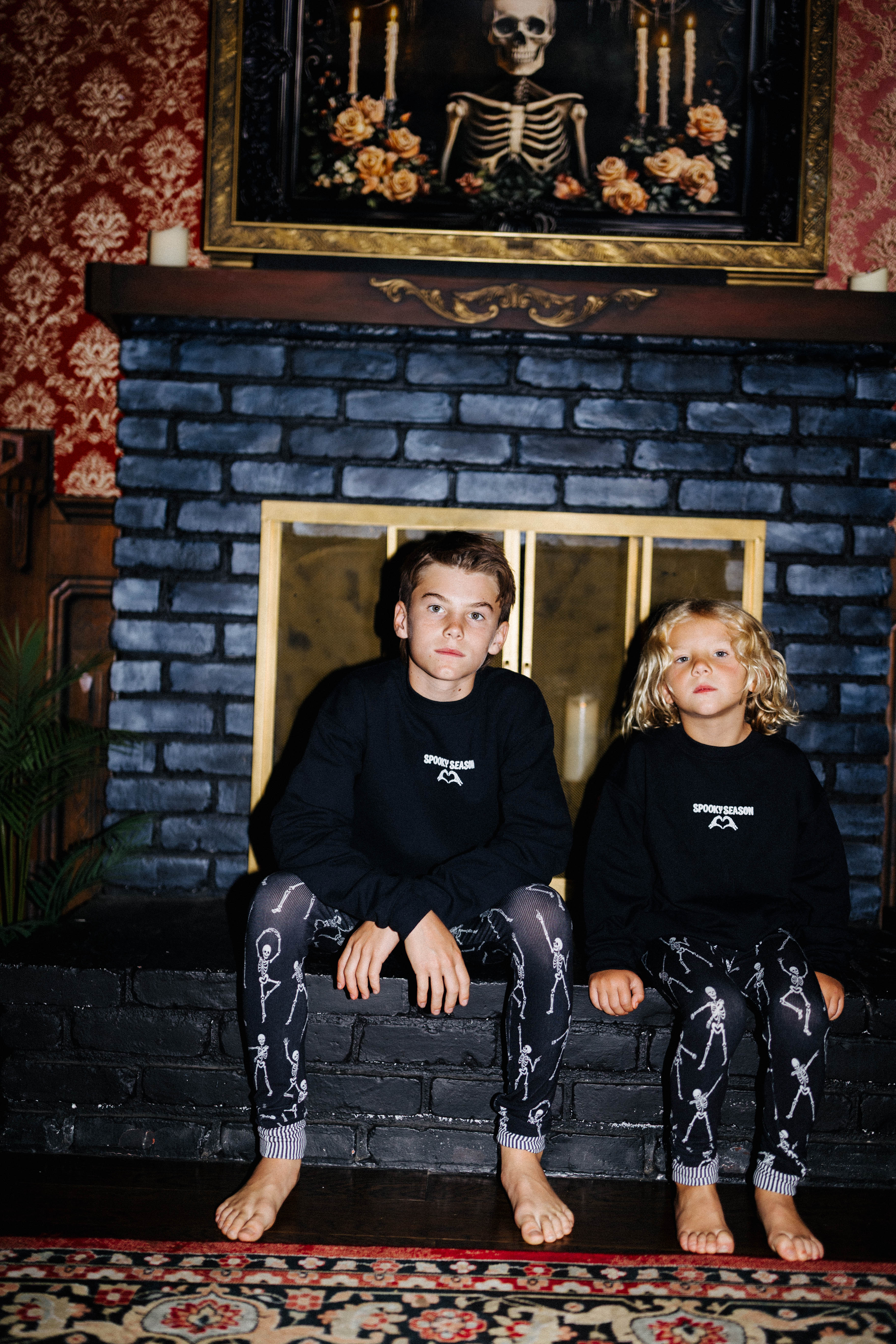 Two children wearing black Spooky Season sweatshirts by a fireplace