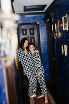 A woman and a young boy wearing matching black and tan checkered pajamas pose together in a dimly lit hallway with blue patterned wallpaper.