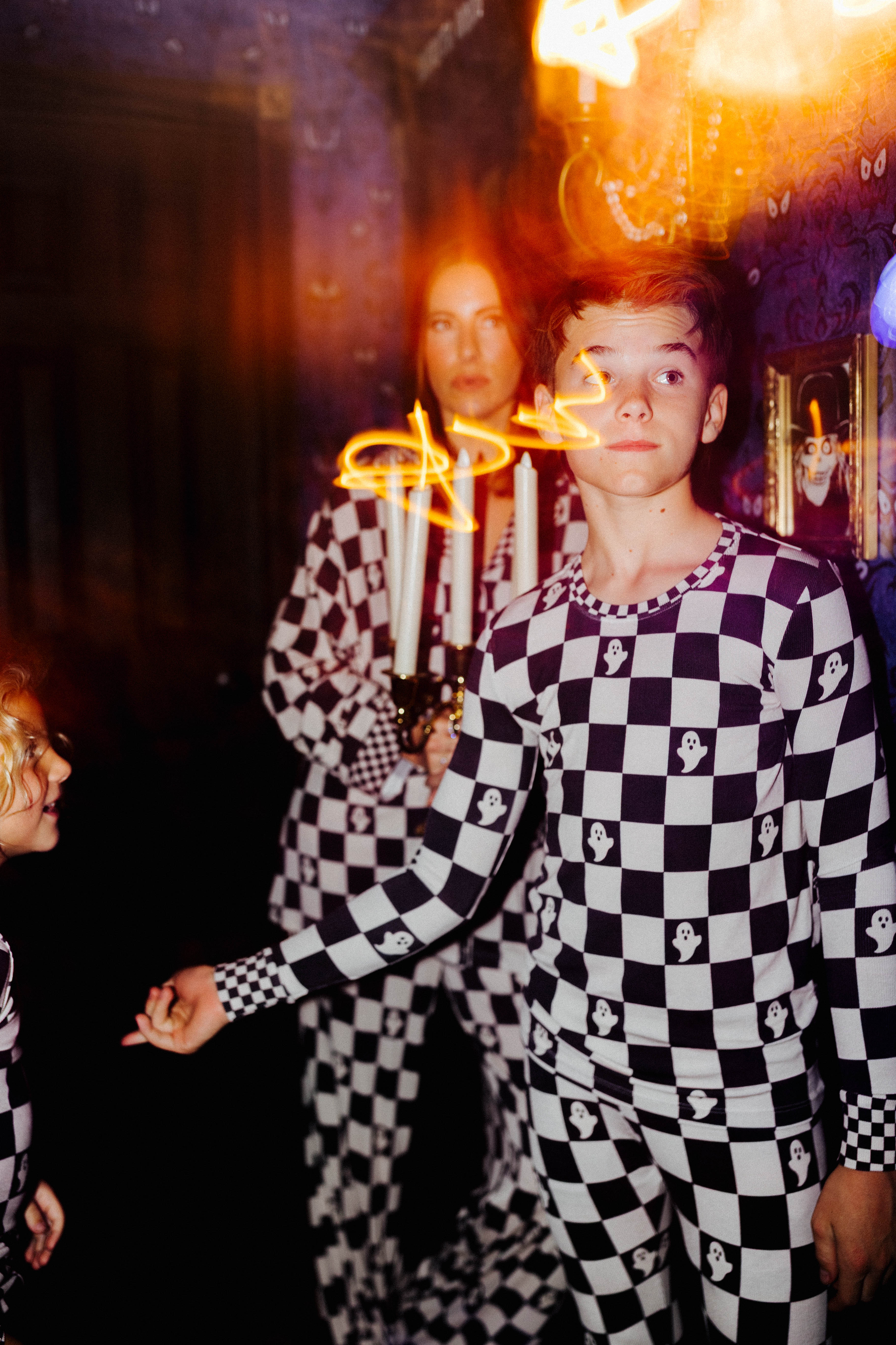 A person in a black-and-tan checkered pajama set stands in a dimly lit room with another person in a matching set behind them, and a bright orange light or flame-like effect across the center.