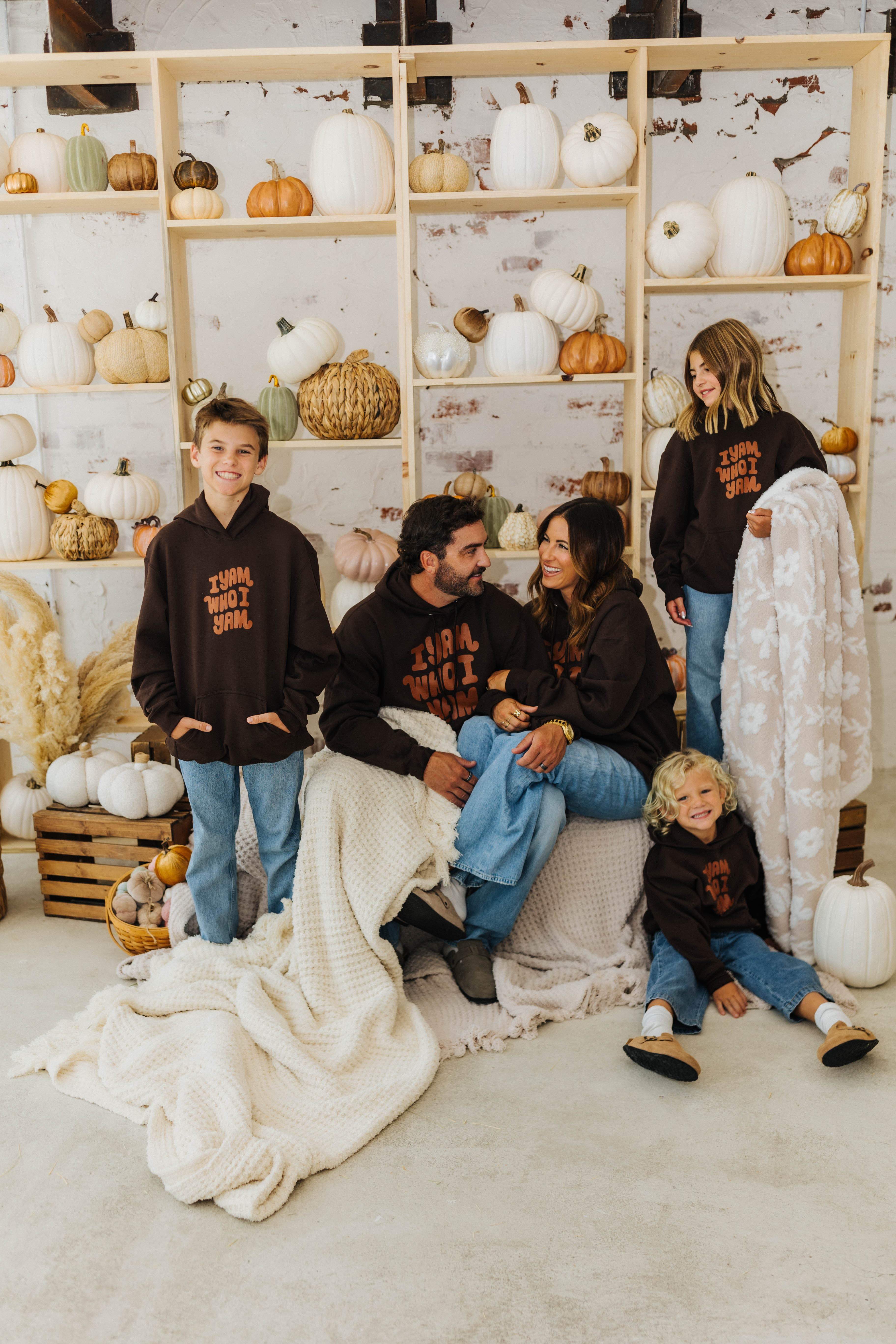 family smiling at each other sitting and standing on hay with waffle blankets and holding pumpkin blanket with pumpkin decorations in the background 
