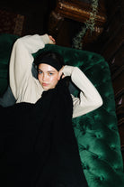 A model wearing a cream long-sleeve top, black headband, and black knit sweater reclines on a dark green tufted velvet sofa in a dimly lit room.