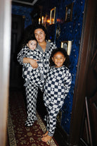 Three people wearing matching black and tan checkered pajamas pose together in a dimly lit hallway with blue patterned wallpaper and a red carpet.