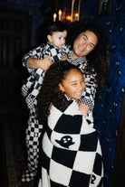 A woman and two children wearing matching sand-and-white checkered cozy pajamas or blankets in a dimly lit room with blue patterned wallpaper and warm candlelight.
