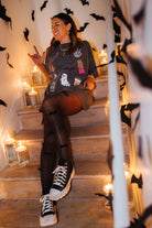 model krista sitting on the staircase wearing hot ghoul halloween tee with halloween graphics and candles and bat decorations in the background 
