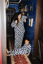 A woman and a child wearing matching black and white checkered pajamas pose in a dark, vintage-style room with blue patterned wallpaper and a red rug.