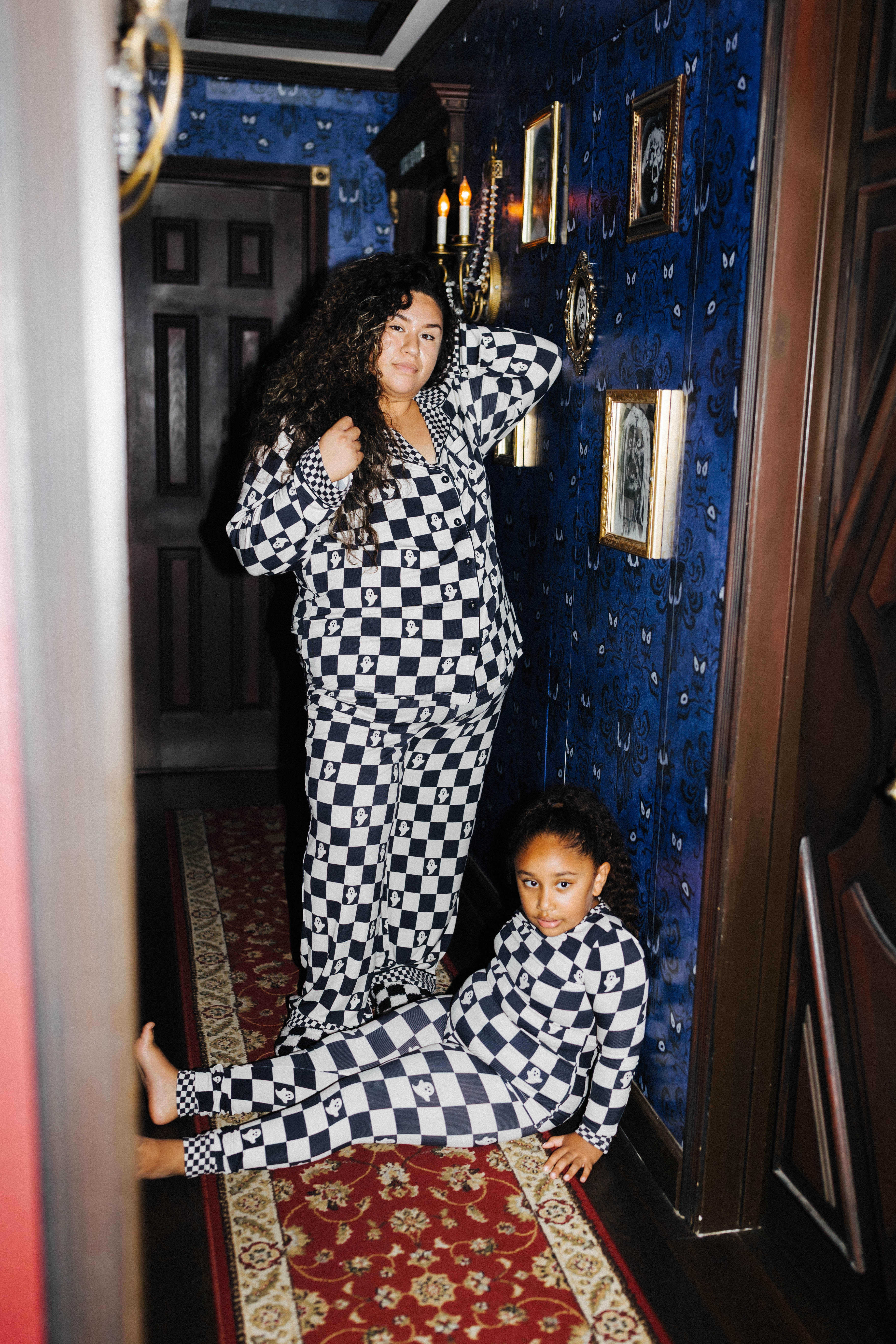 A woman and a child wearing matching black and white checkered pajamas pose in a dark, vintage-style room with blue patterned wallpaper and a red rug.