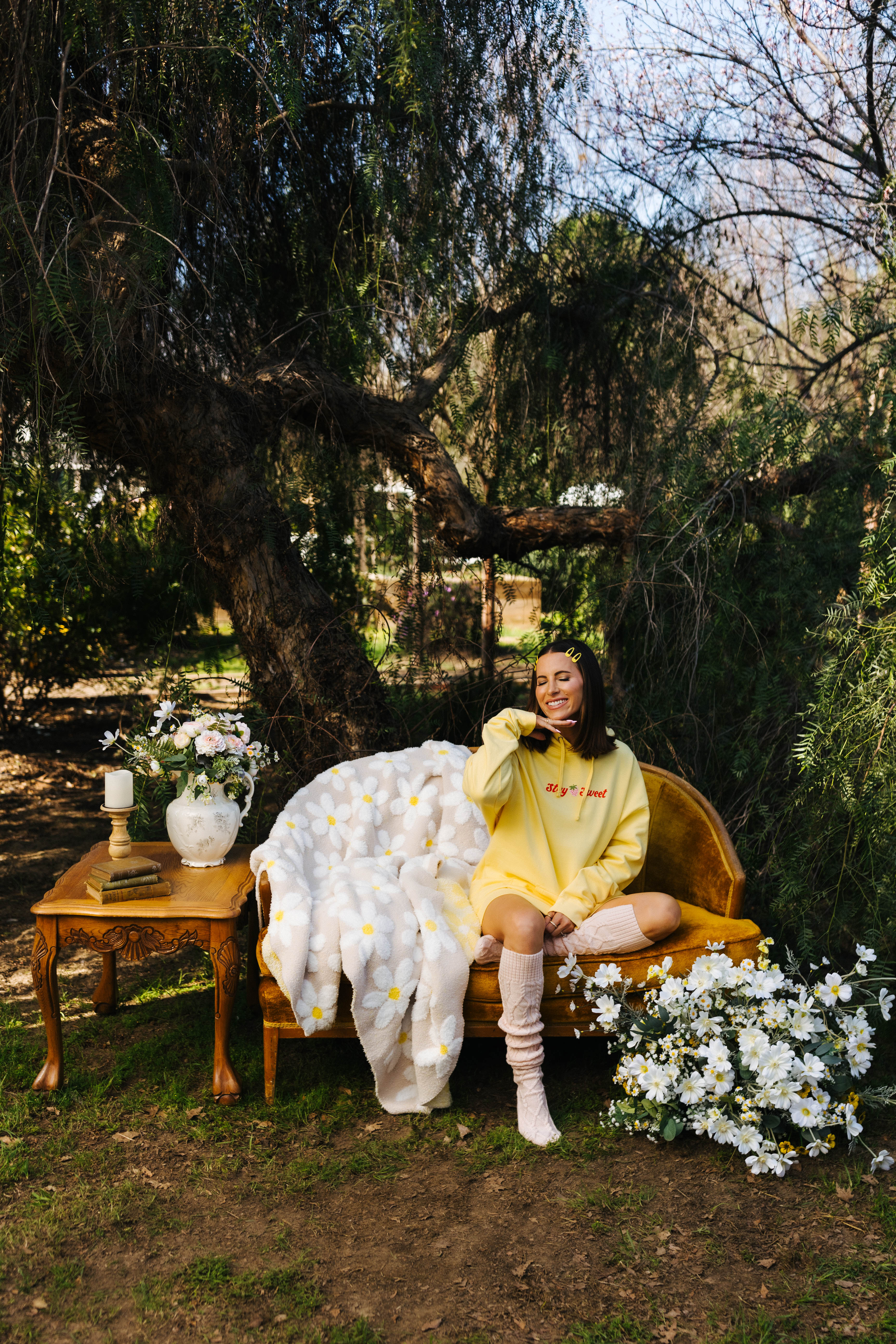 Person in a yellow outfit sitting on a chair with a sand b
lanket and flowers around, under a tree.