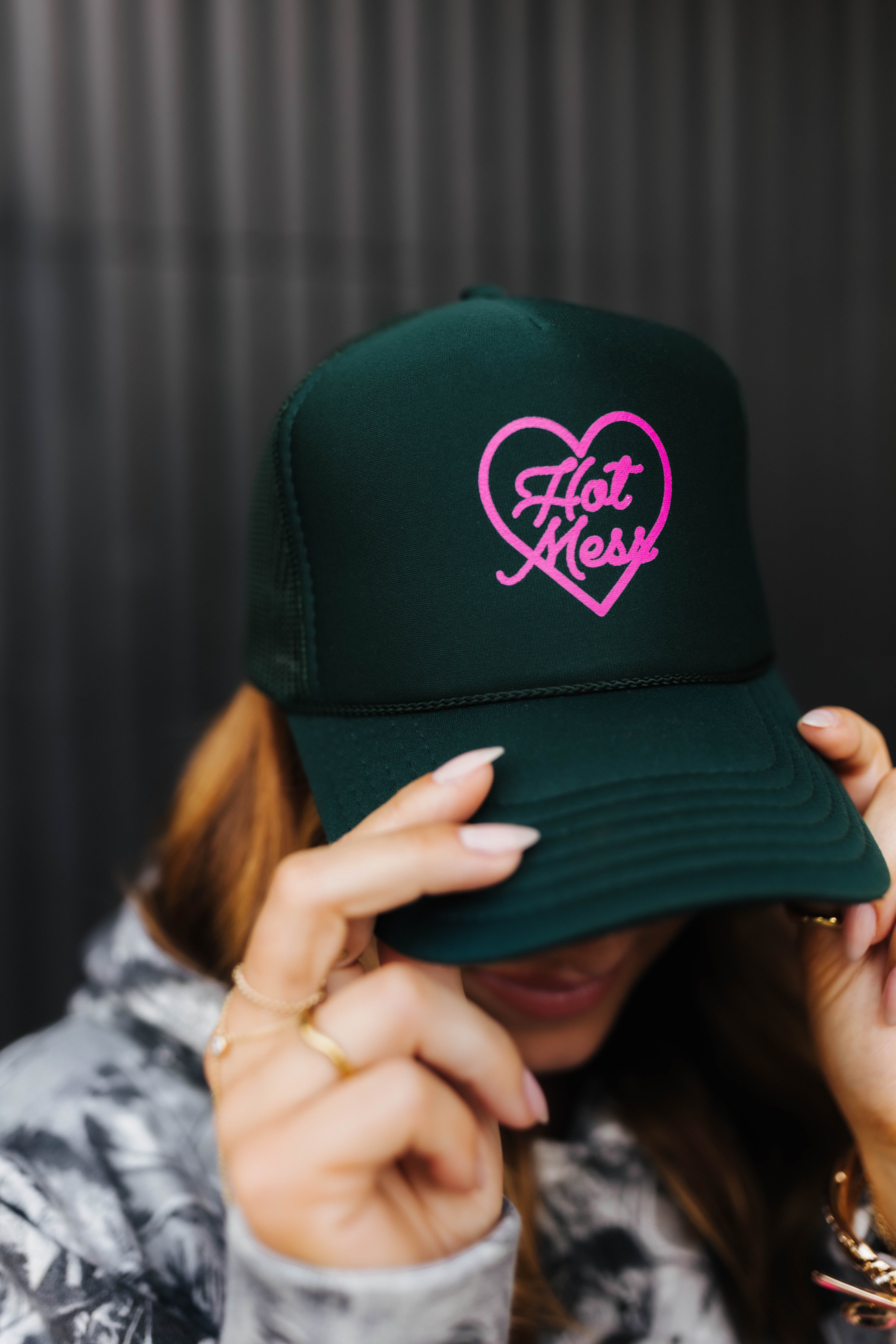 model holding dark green hot mess at heart hat against black wall 