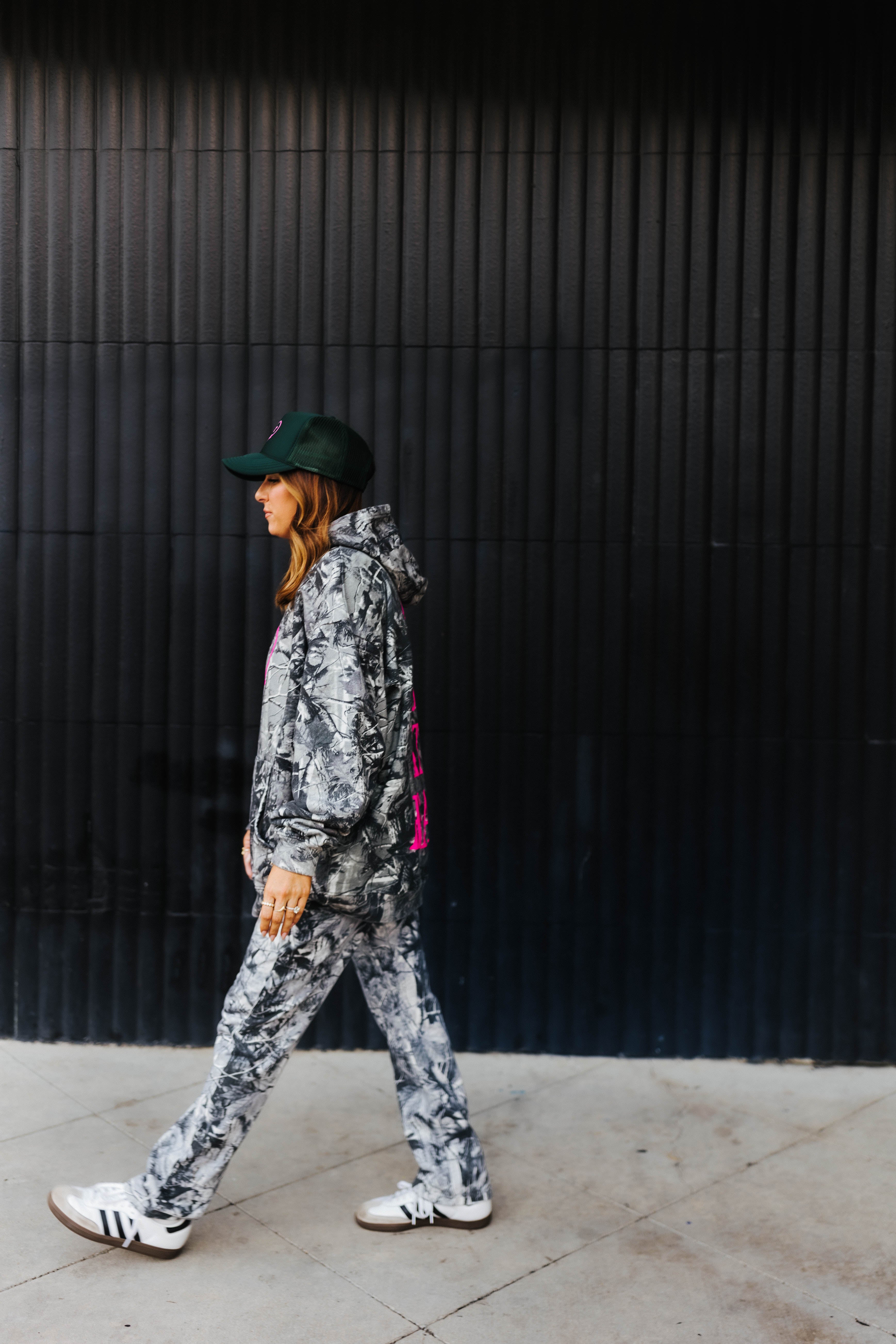 model walking away showing hot mess at heart camo hoodie and hot mess at heart hat against black wall 