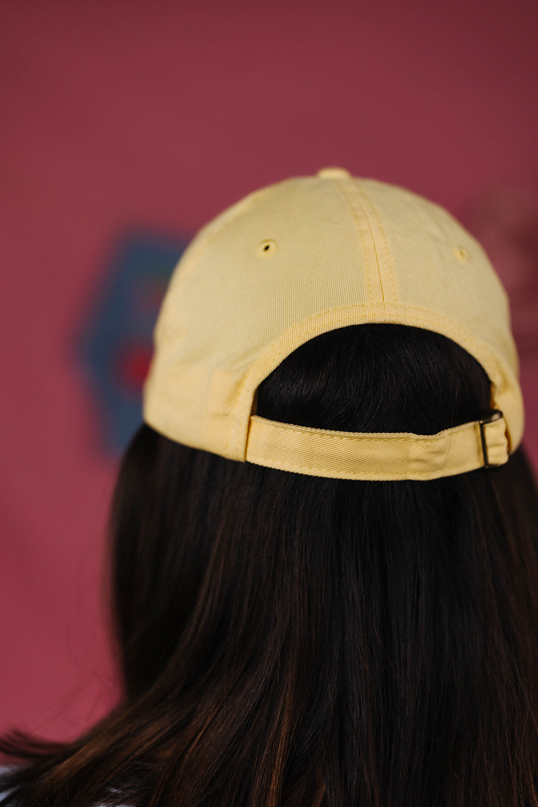 back of yellow cap worn by a person with long dark hair against a blurred background