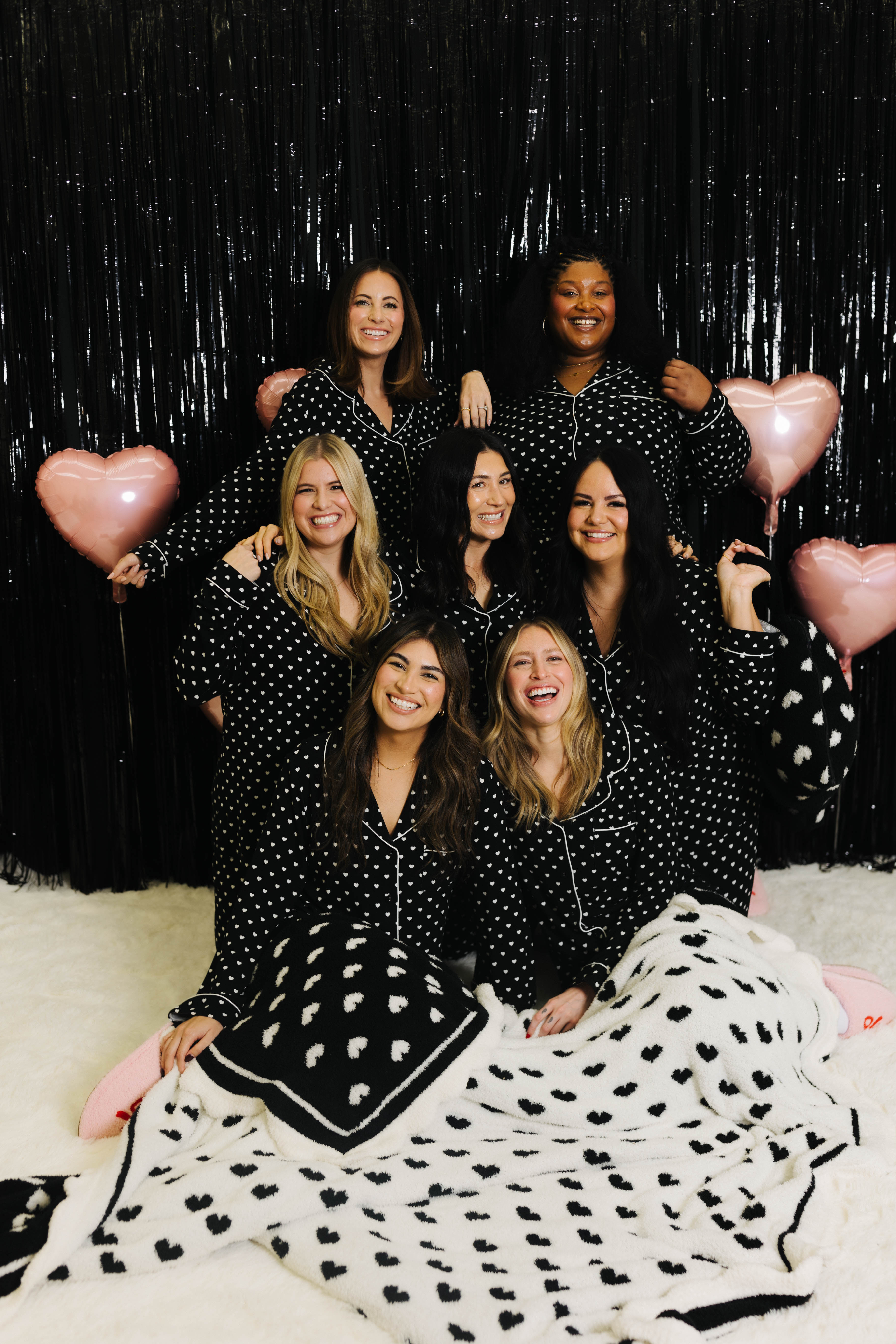 Group of women in matching polka dot outfits with pink balloons against a black glittery background.