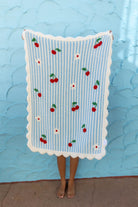 A person holding up a light blue and white striped blanket with a scalloped white border and embroidered red cherries, standing against a textured light blue wall.