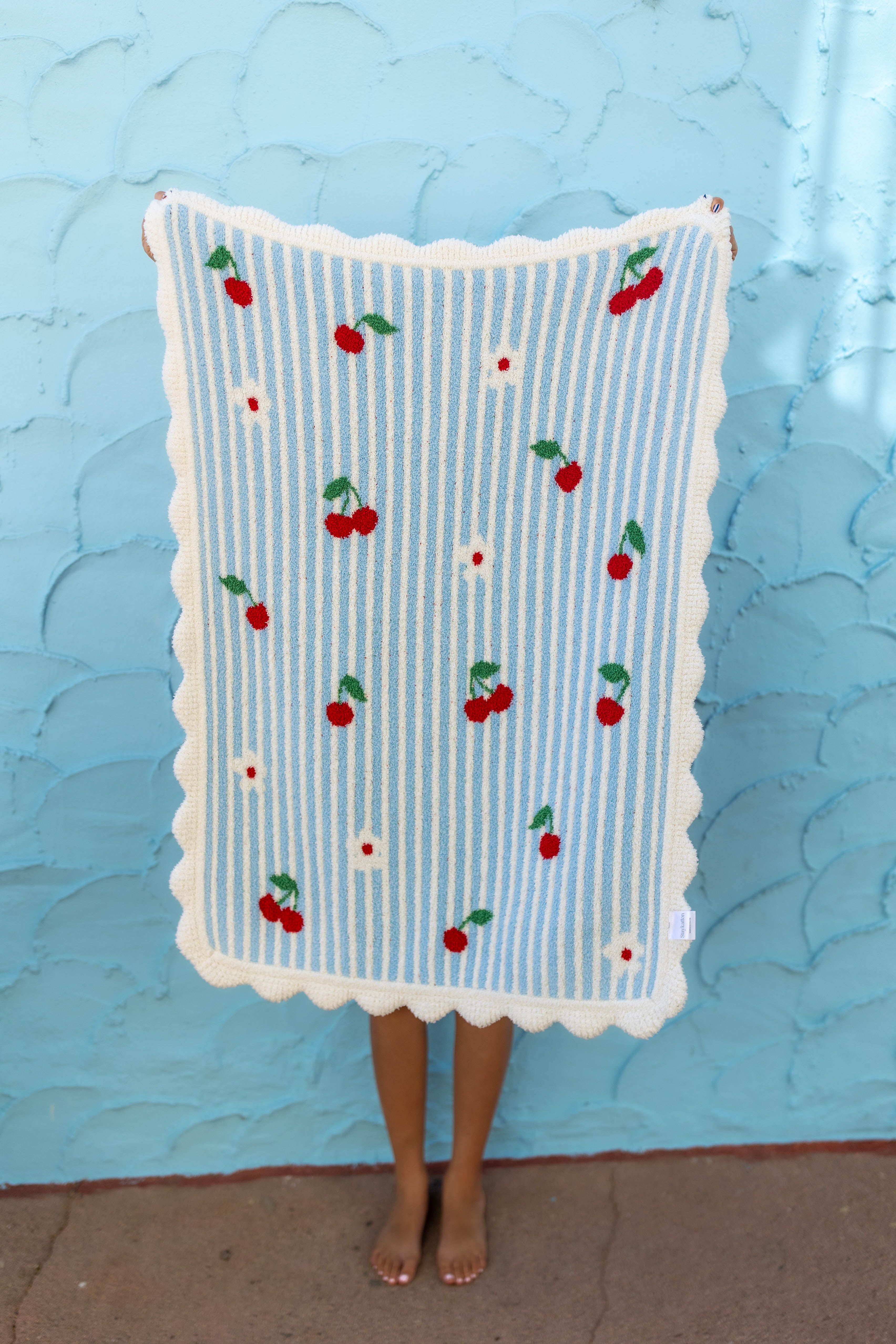 A person holding up a light blue and white striped blanket with a scalloped white border and embroidered red cherries, standing against a textured light blue wall.