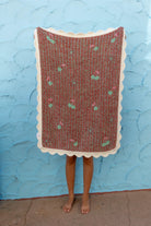 A person standing barefoot in front of a textured light blue wall, holding up a rectangular woven blanket with a scalloped cream border. 