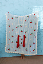 A light blue and white striped blanket with a cherry and daisy pattern, featuring a front pocket with red drawstrings, held up by a person standing against a textured light blue wall.