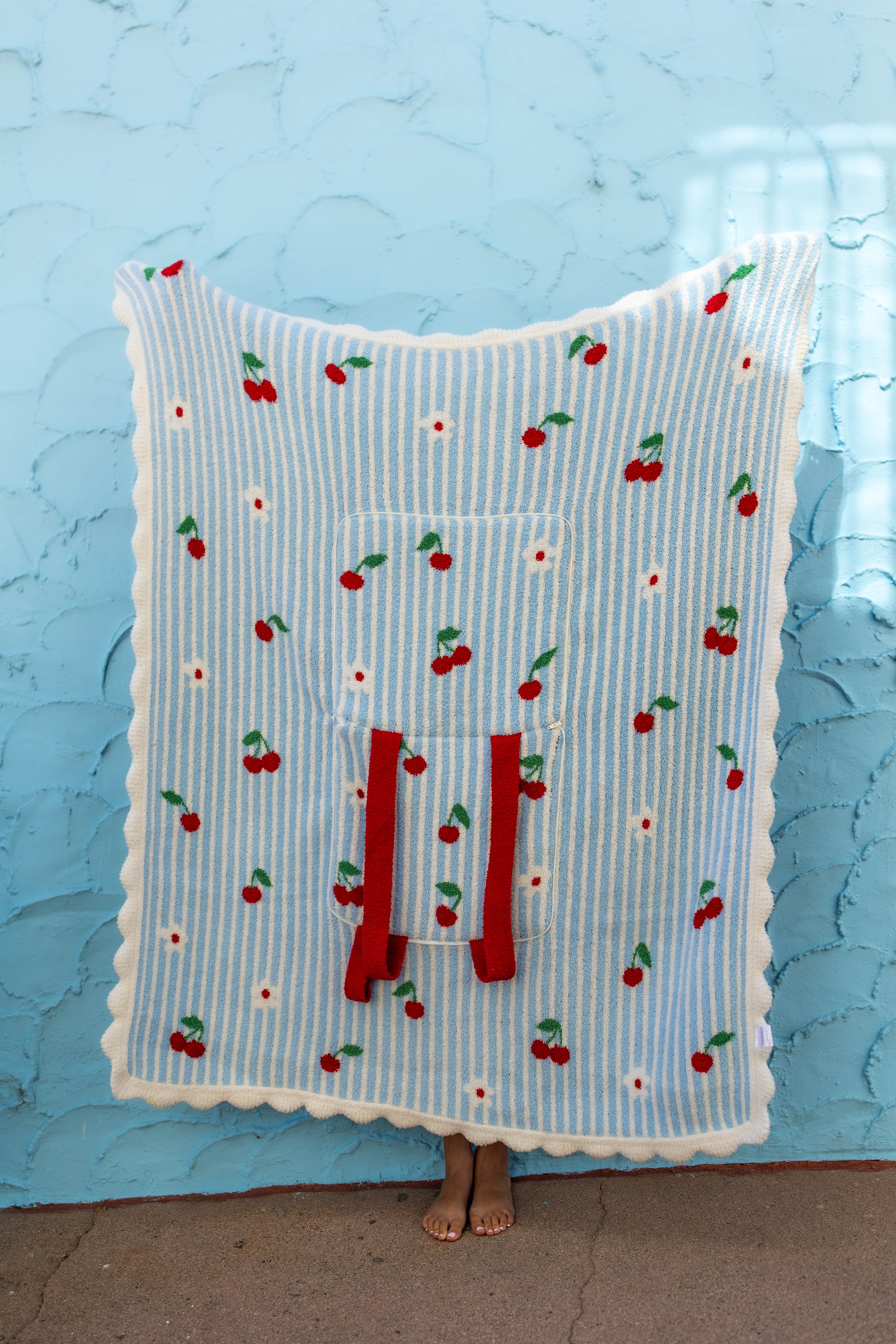 A light blue and white striped blanket with a cherry and daisy pattern, featuring a front pocket with red drawstrings, held up by a person standing against a textured light blue wall.