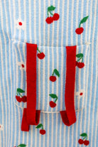 Close-up of a light blue and white striped fabric with embroidered red cherries and green leaves, featuring a front pocket with red straps.