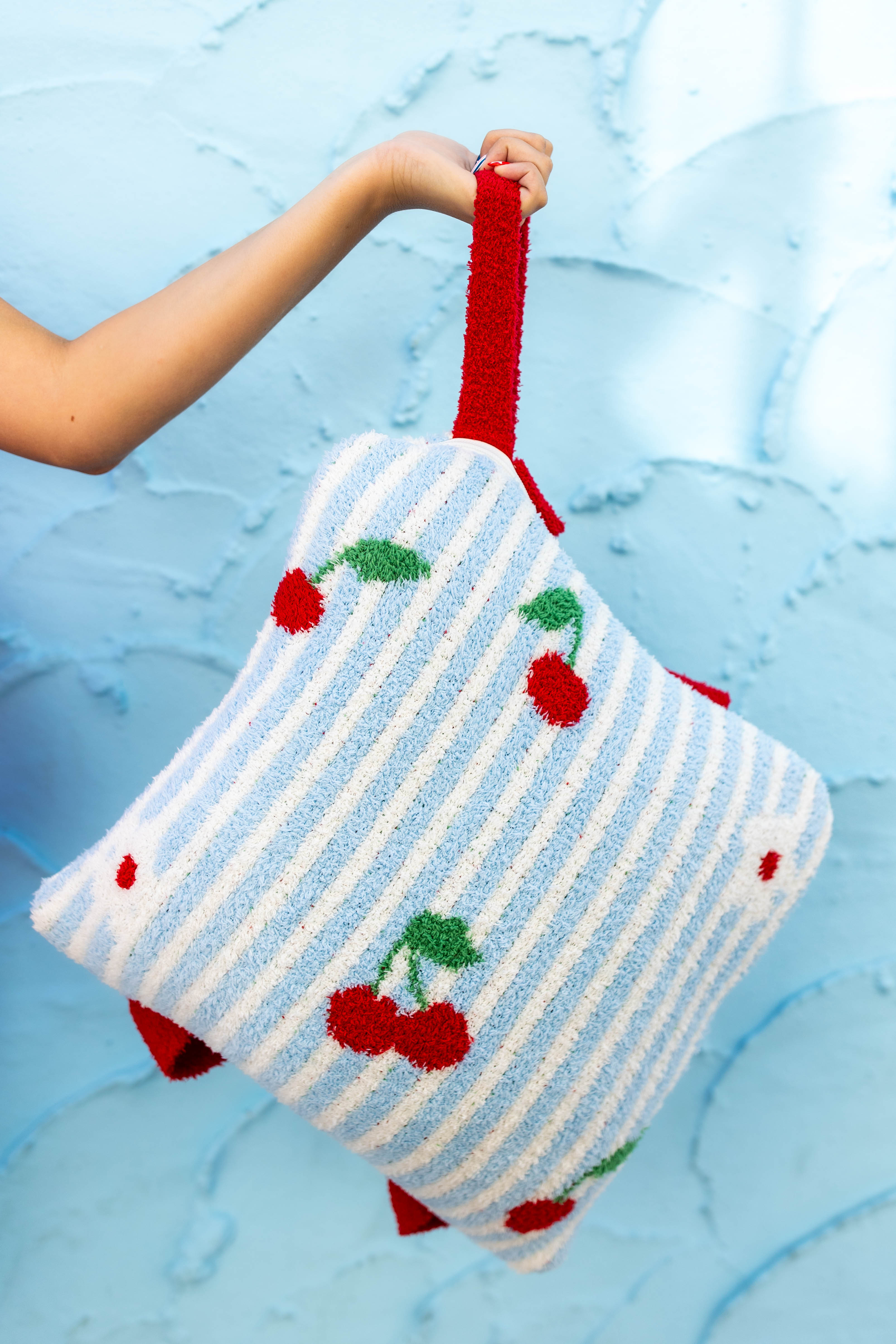 A fuzzy knit quillow with a white and light blue vertical stripe pattern and red cherries with green leaves, held by a person's hand against a textured light blue background.