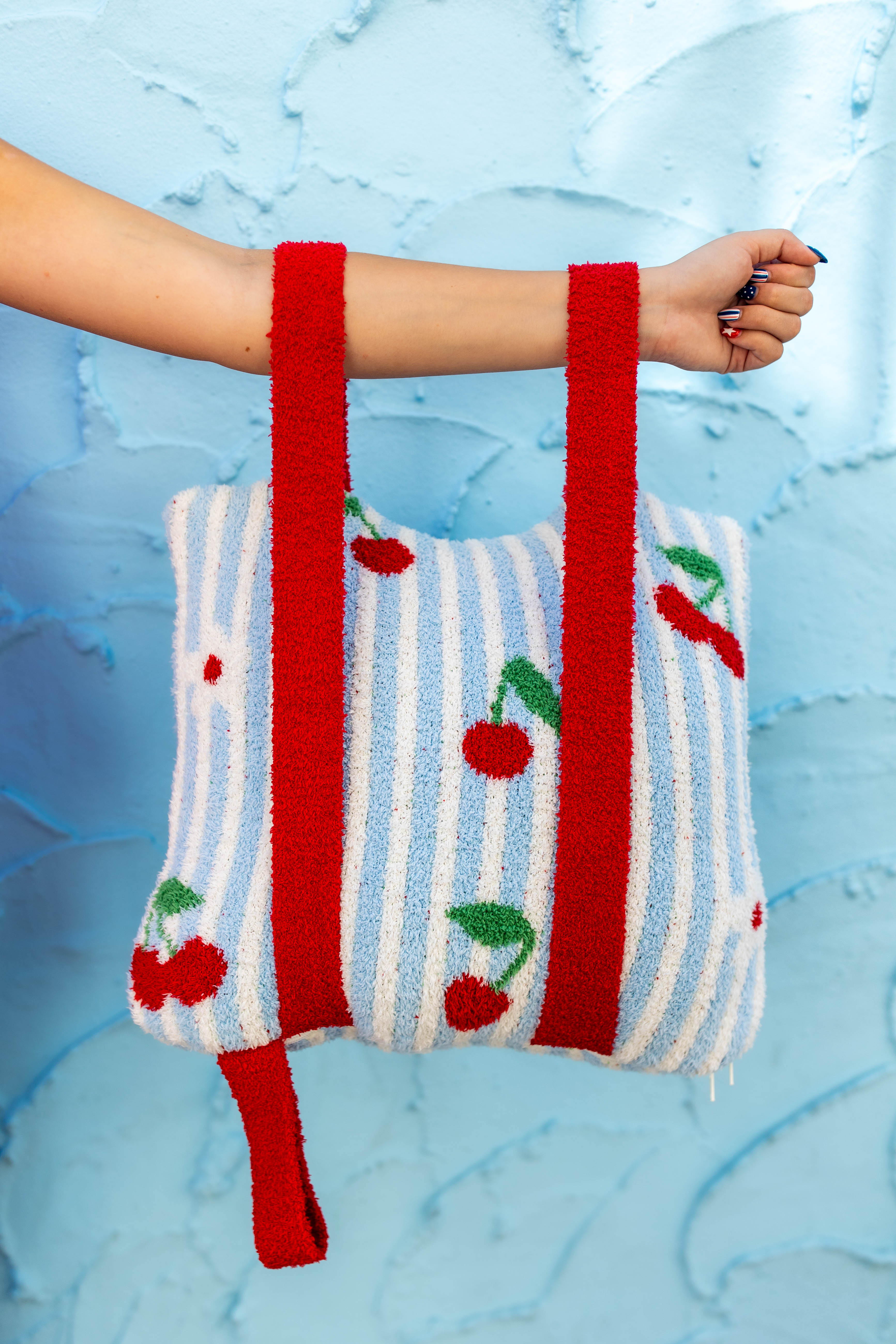 A fuzzy knit quillow with red handles and a white and light blue vertical stripe pattern decorated with red cherries with green leaves, held by a person's arm against a textured light blue background.