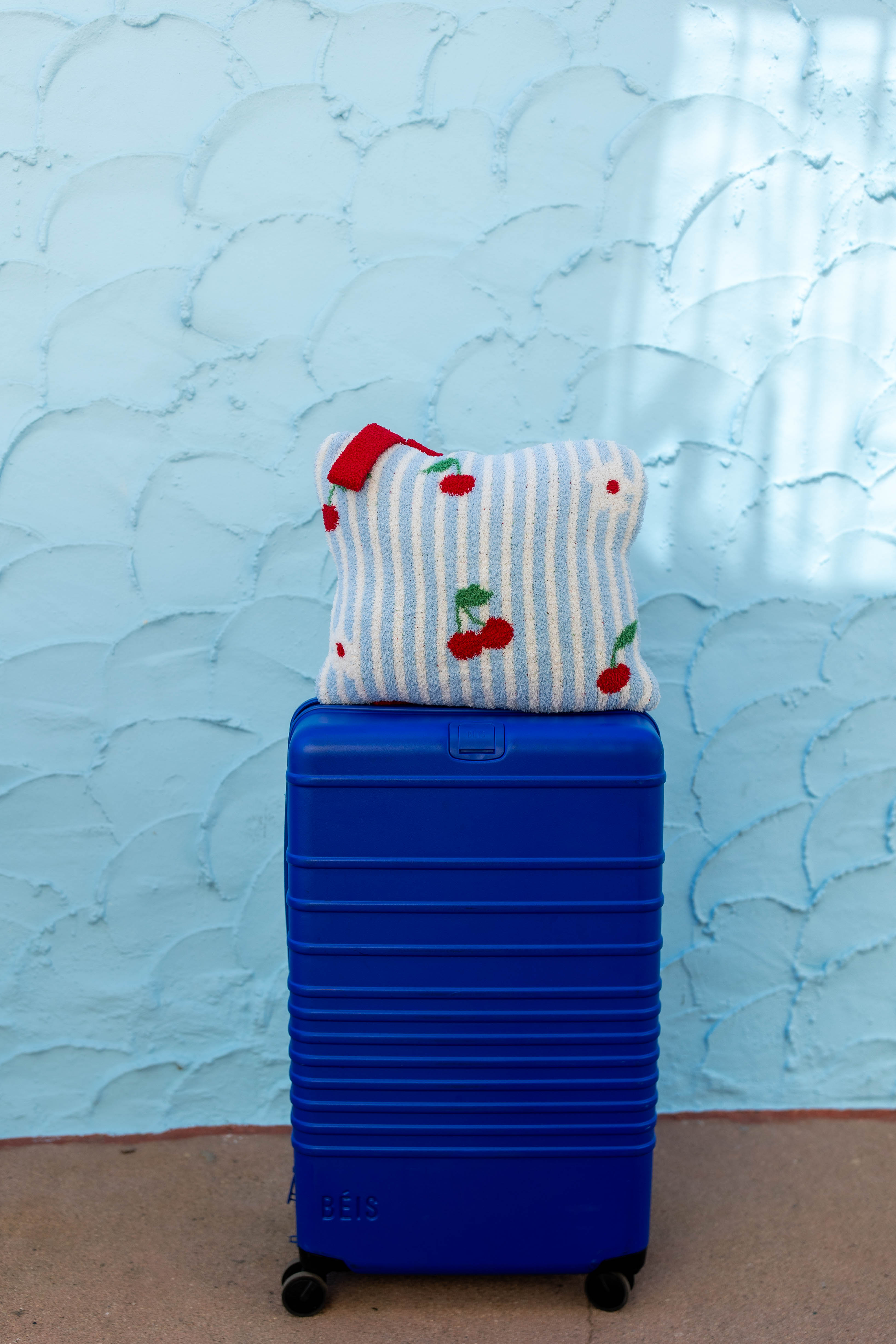 A bright blue hard-shell suitcase standing upright on a tan floor against a light blue textured wall, with a striped and cherry-patterned quillow resting on top.