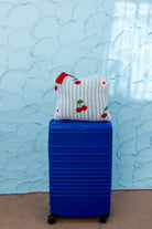 A bright blue hard-shell suitcase standing upright on a tan floor against a light blue textured wall, with a striped and cherry-patterned quillow resting on top.
