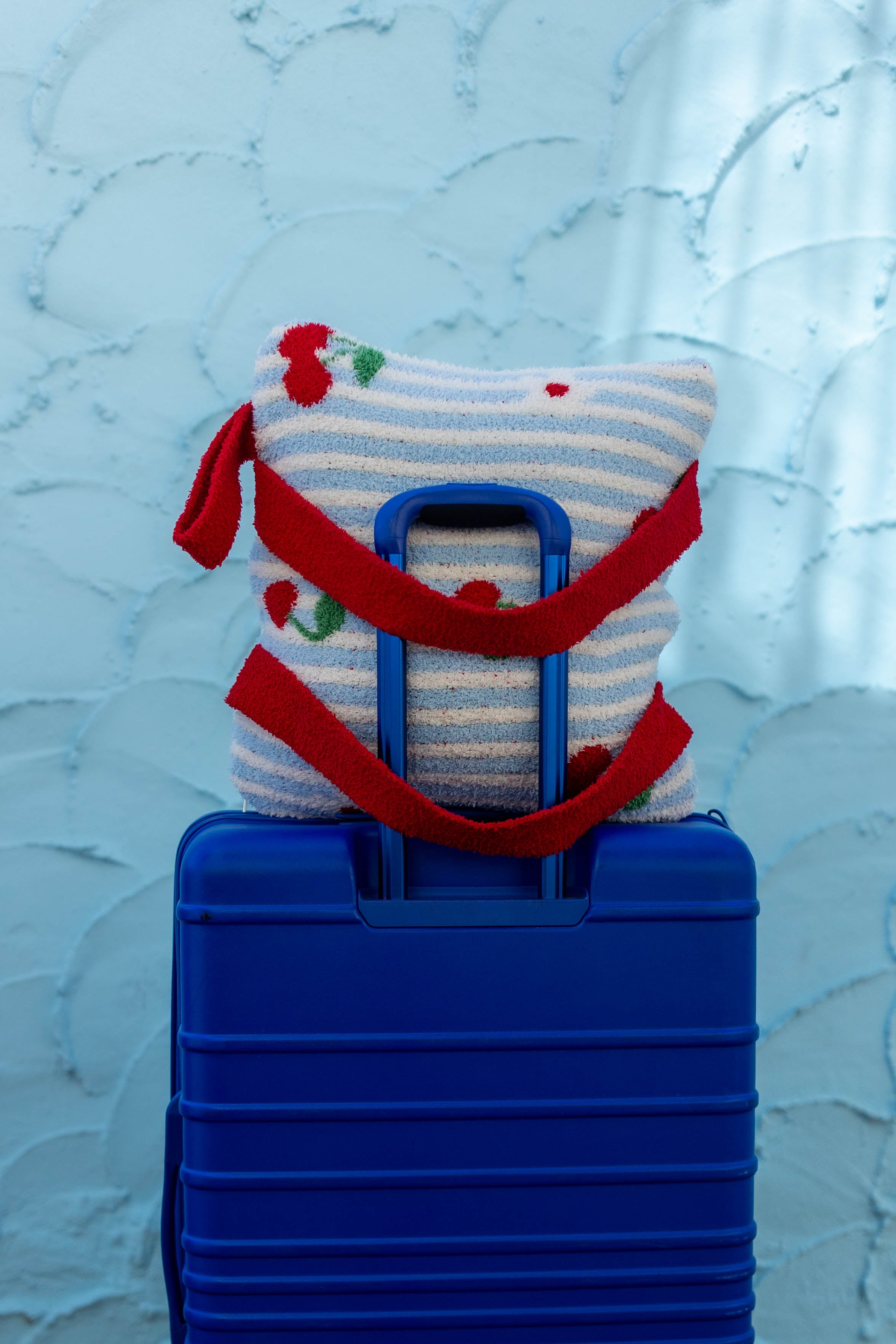 A blue hard-shell suitcase with the handle extended, topped with a striped white and light blue travel quillow and wrapped in a red scarf, set against a textured light blue wall.