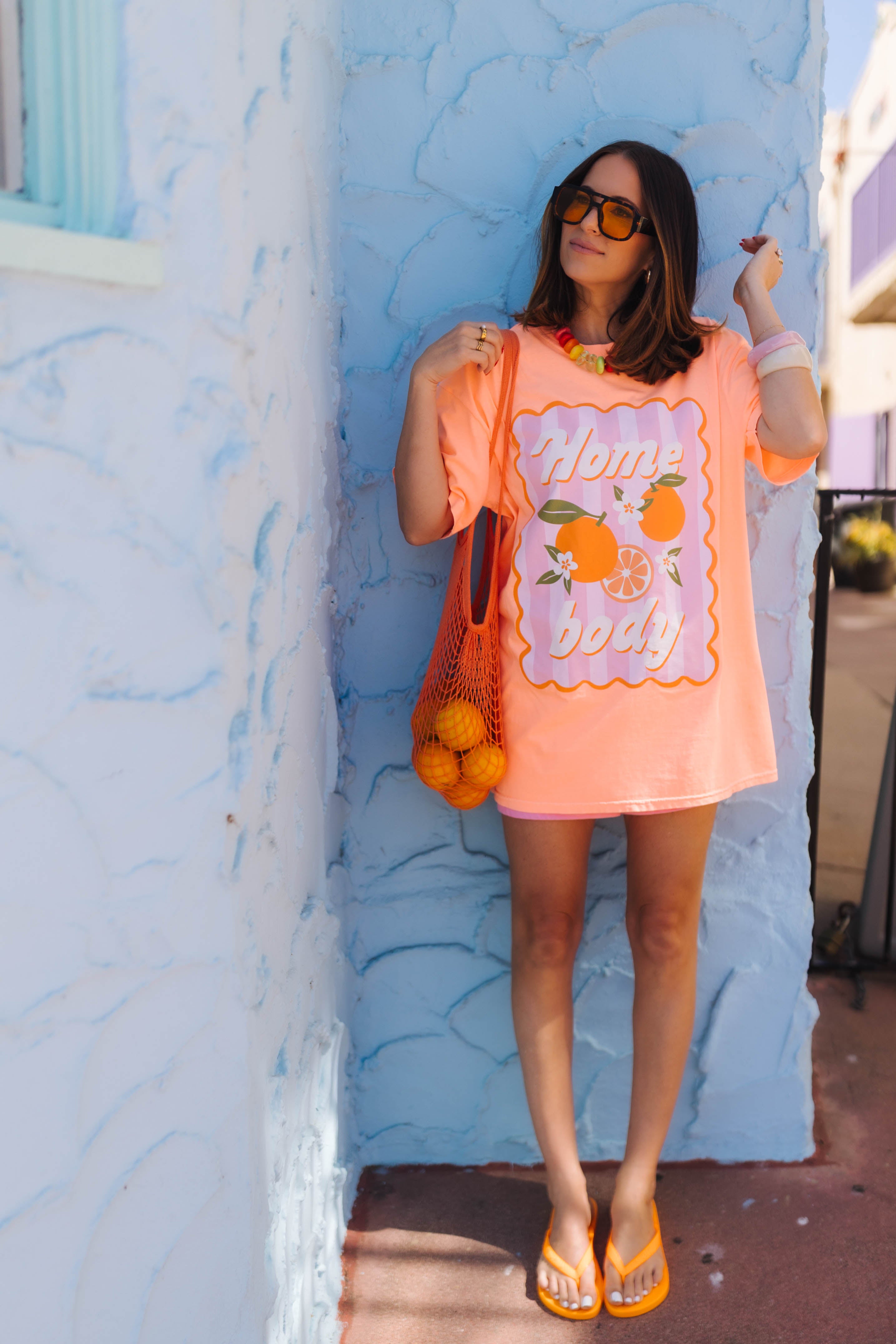 A woman stands outdoors against a light blue textured wall wearing an oversized orange graphic T-shirt with orange fruit illustrations and the words 'Home body,' along with orange flip-flops and a mesh tote bag filled with oranges.