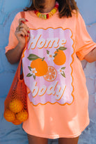 A person wearing an orange oversized graphic T-shirt with the words 'Home body' in script over orange and citrus illustrations. The person is holding an orange mesh tote bag and standing against a light blue textured wall.
