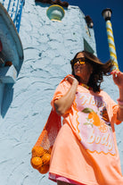 A woman wearing a bright orange graphic t-shirt and yellow-tinted sunglasses, carrying an orange mesh bag of oranges against a light blue textured stucco wall.