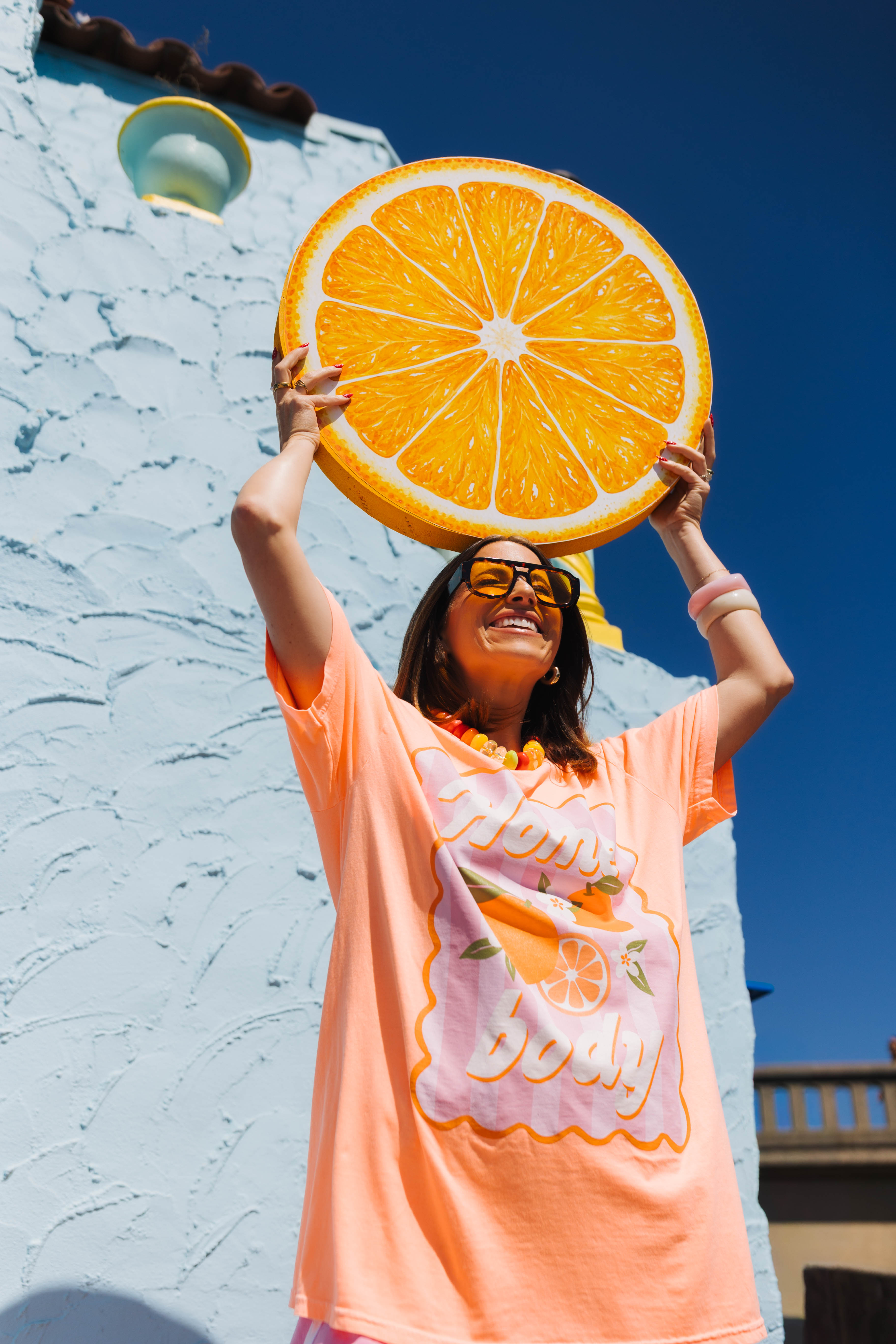 A woman wearing an orange graphic t-shirt and glasses holds a giant orange slice prop above her head, posing in front of a white stucco building under a clear blue sky.