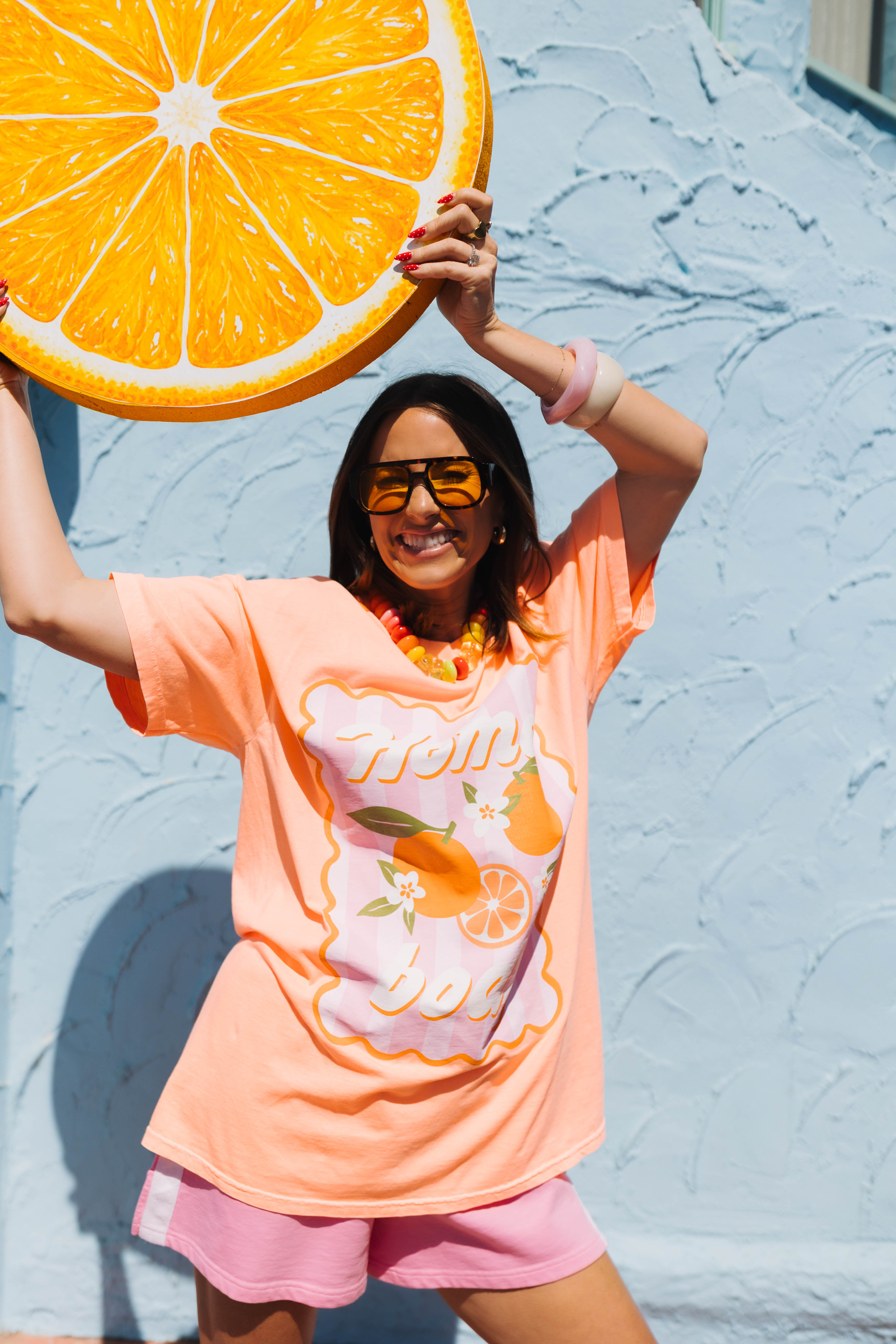 A woman wearing an orange graphic t-shirt with an orange and pineapple print, pink shorts, and yellow sunglasses, holding a giant orange slice prop against a textured light blue wall.
