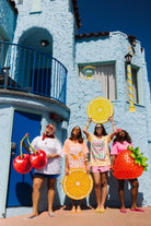 Four women stand in front of a light blue Mediterranean-style building, each holding a large fruit prop: two red cherries, one lemon, one range slice, and one strawberry. 