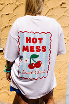A person viewed from behind wearing an oversized white graphic T-shirt with a large retro-style red, blue, and cherry print on the back, standing against a textured yellow wall.