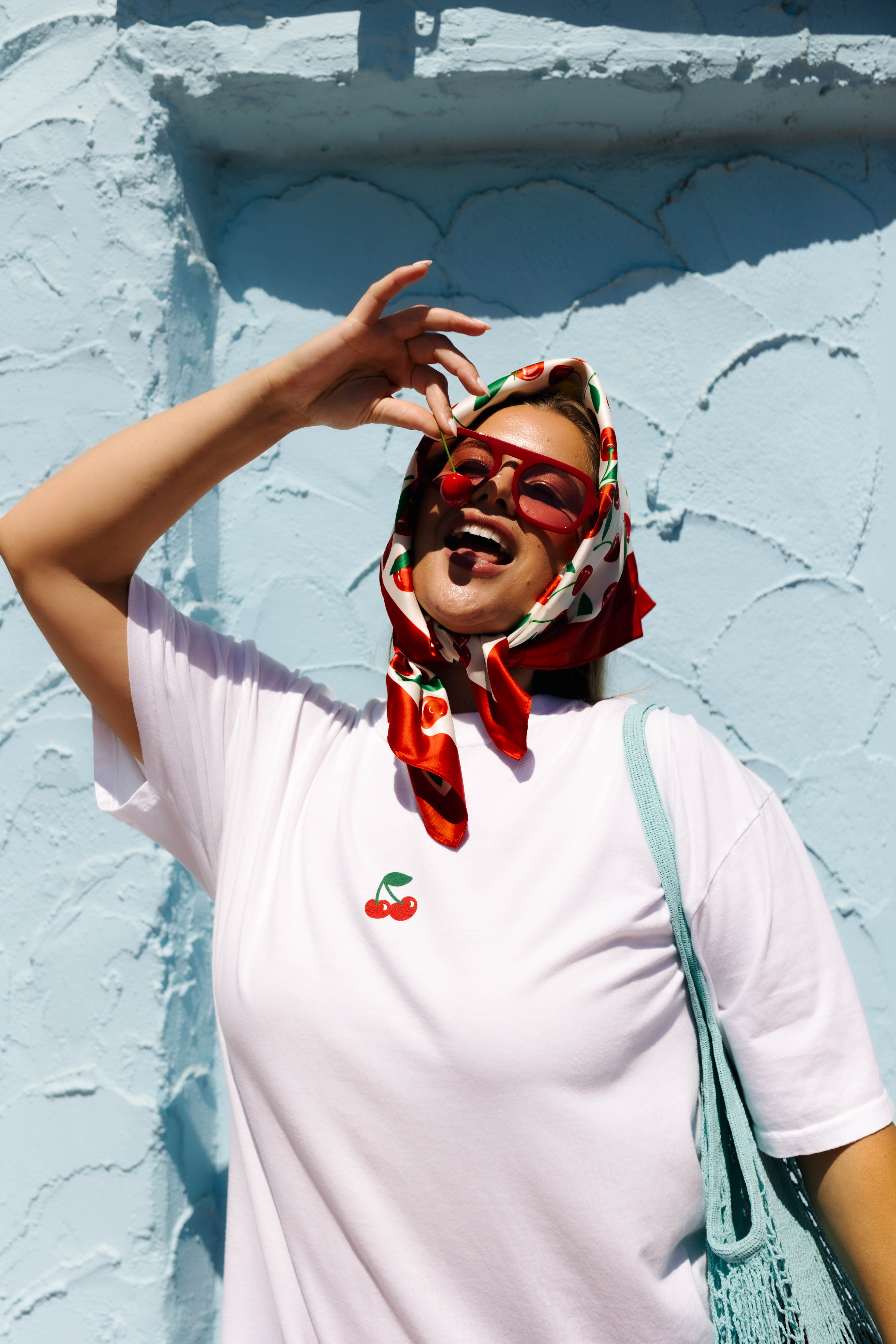 A woman wearing a white t-shirt with a small cherry graphic on the chest, a red patterned headscarf, and red sunglasses, posing against a light blue textured wall.