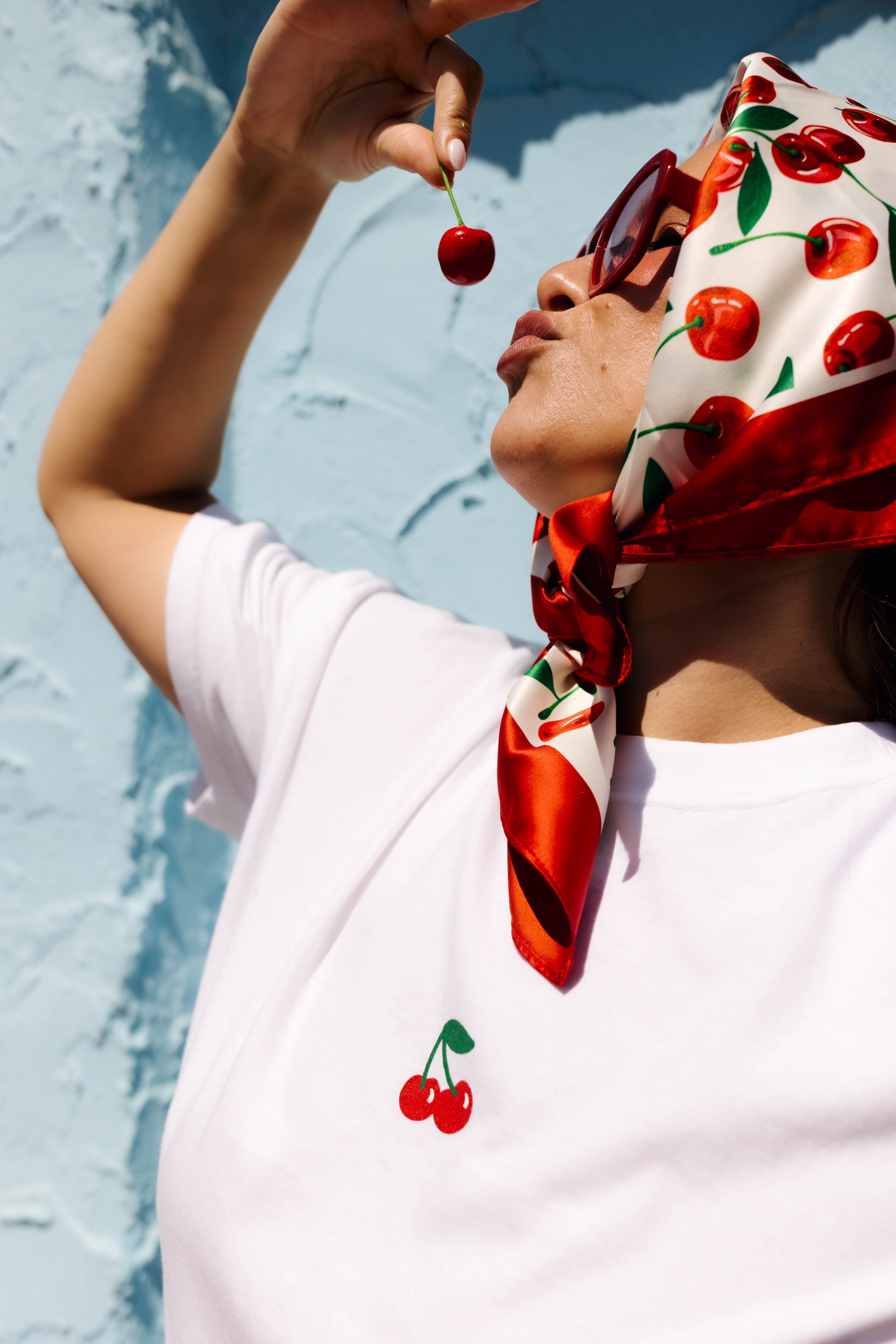 A woman wearing a white t-shirt with a red cherry graphic on the chest, a red and white cherry-patterned silk headscarf, and dark sunglasses, holding a cherry near her mouth against a textured light blue wall.
